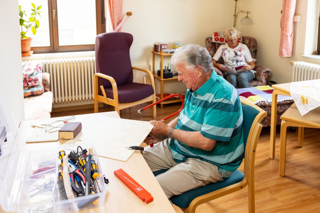 Ein alter Mann macht Handarbeiten und eine alte Frau liest ein Buch im Hintergrund